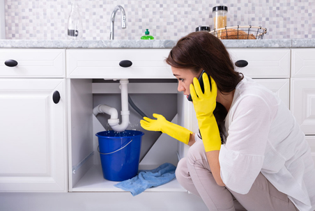 A woman in yellow gloves is on the phone, looking at a leaky pipe